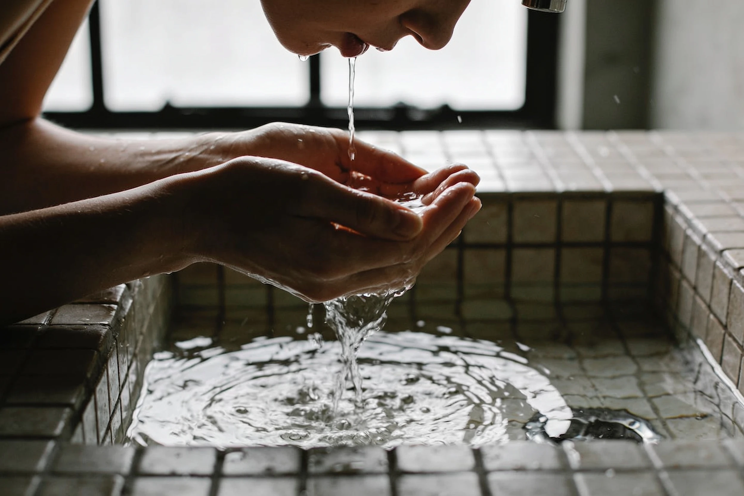 Hands cupping water under a tap while rinsing the face at a stone basin