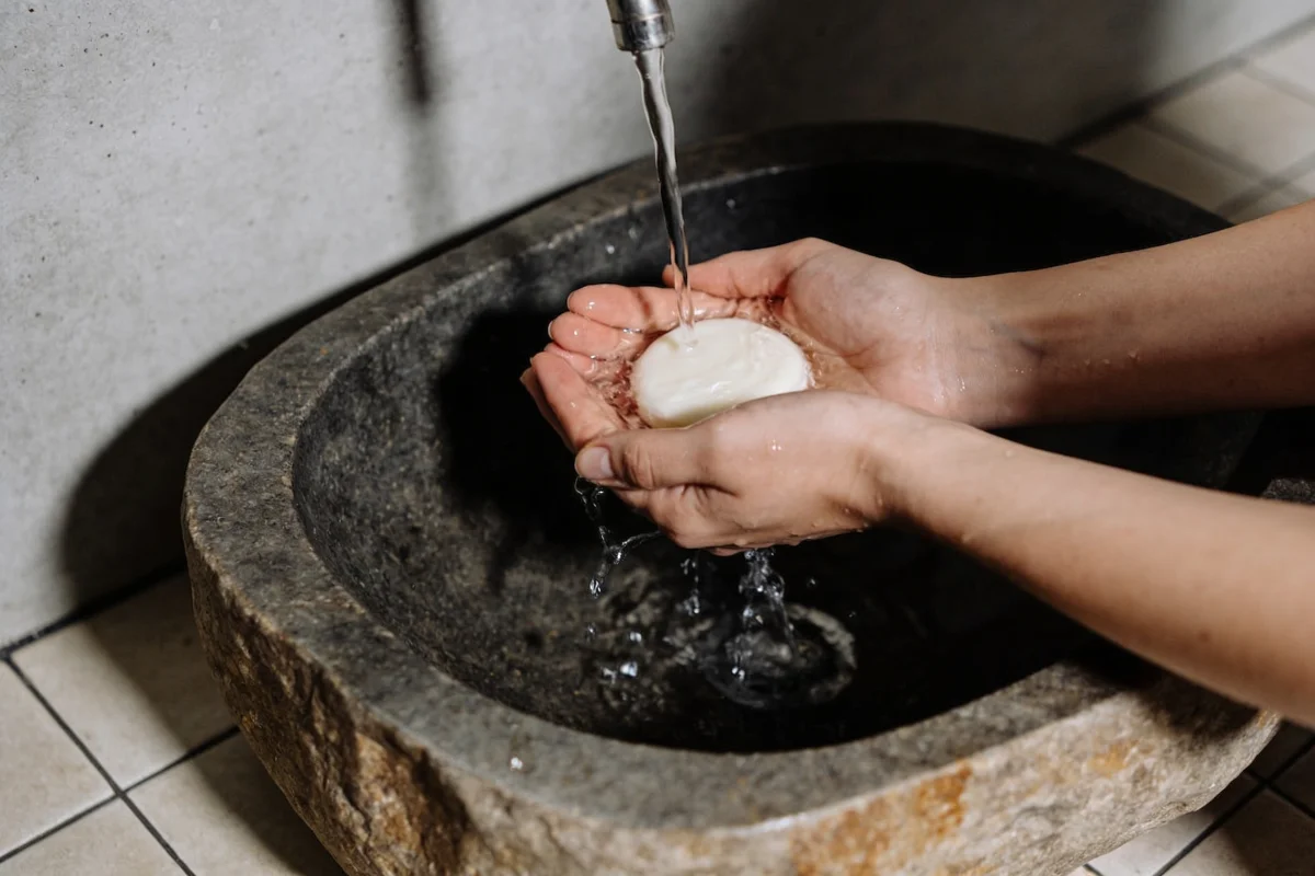 Hands cupping water and a bar of cleanser at a stone basin in soft light, how to wash your face